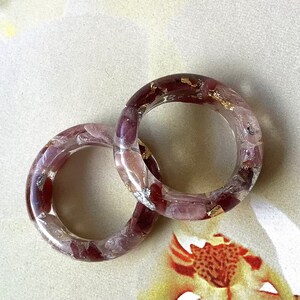 May include: Two clear resin rings with embedded red and pink stones and gold leaf accents. The rings are circular and have a polished finish, showcasing the colorful inclusions. The rings are likely handmade and are suitable for everyday wear.