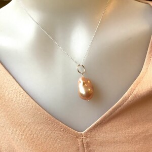 May include: A silver chain necklace with a large, irregularly shaped, peach-colored pearl pendant. The pearl is suspended from a small silver ring. The necklace is displayed on a mannequin.