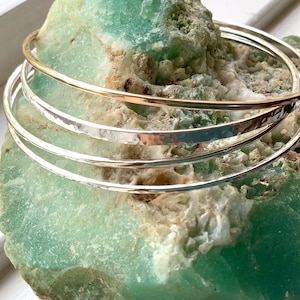 May include: Four metal bangles are shown. One is gold-toned, the others silver-toned. They rest on a light green, textured surface. Three silver bangles have a hammered texture. The bangles are simple and elegant.