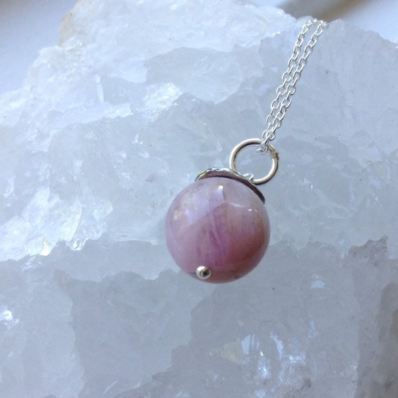 Op de afbeelding: Zilveren ketting met een ronde, roze edelsteen hanger. De hanger heeft een zilveren dop en een kleine zilveren kraal aan de onderkant. De edelsteen heeft een gemarmerd uiterlijk.