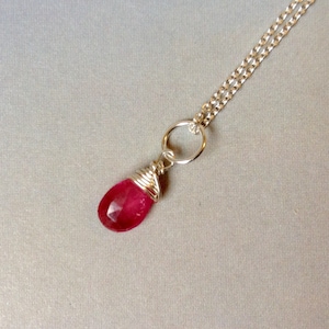 Peut inclure: Un collier en argent avec un pendentif en forme de goutte d'eau rouge rubis facetté. La pierre est enveloppée de fil d'argent et suspendue à une chaîne en argent. Le collier est présenté sur un fond gris neutre.