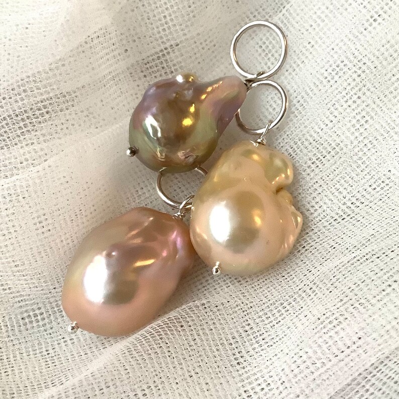 May include: Three iridescent baroque pearls with silver-tone metal loops. The pearls have a lustrous sheen, displaying colors of pink, gold, and lavender. The pearls are irregularly shaped, with a natural, organic appearance. The background is a white, textured fabric.