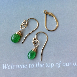 May include: Gold-coloured earrings featuring teardrop-shaped green gemstones. The gemstones are wire-wrapped and suspended from gold-coloured ear wires. A gold-coloured earring clasp is also visible. The earrings are displayed on a light grey surface.