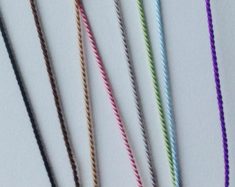 May include: Close-up of several strands of twisted cord in various colors, including black, brown, pink, green, blue, and purple. The cords are arranged diagonally against a white background, showcasing their texture and color variations.