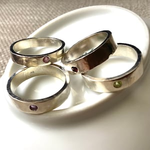 May include: Four silver rings with a hammered texture, each set with a small gemstone in shades of pink and green. The rings are displayed on a white, oval-shaped dish.
