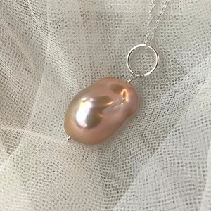 May include: A silver necklace featuring a large, irregularly shaped, peach-colored pearl pendant. The pearl has a lustrous surface with iridescent highlights. The pendant is suspended from a silver chain through a small silver ring.