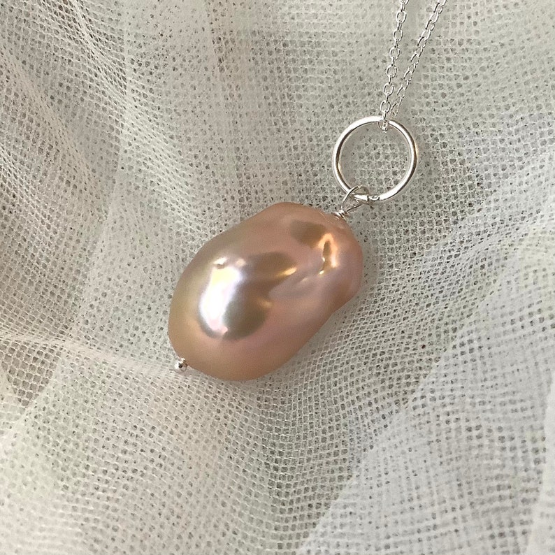 May include: A silver necklace featuring a large, irregularly shaped, peach-colored pearl pendant. The pearl has a lustrous surface with iridescent highlights. The pendant is suspended from a silver chain through a small silver ring.