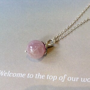 Op de afbeelding: Een delicate zilveren ketting met een ronde, lichtroze edelsteen hanger. De hanger wordt op zijn plaats gehouden door een zilveren zetting. De ketting is dun en de achtergrond is lichtgrijs. De tekst "Welcome to the top of our wo..." is zichtbaar.