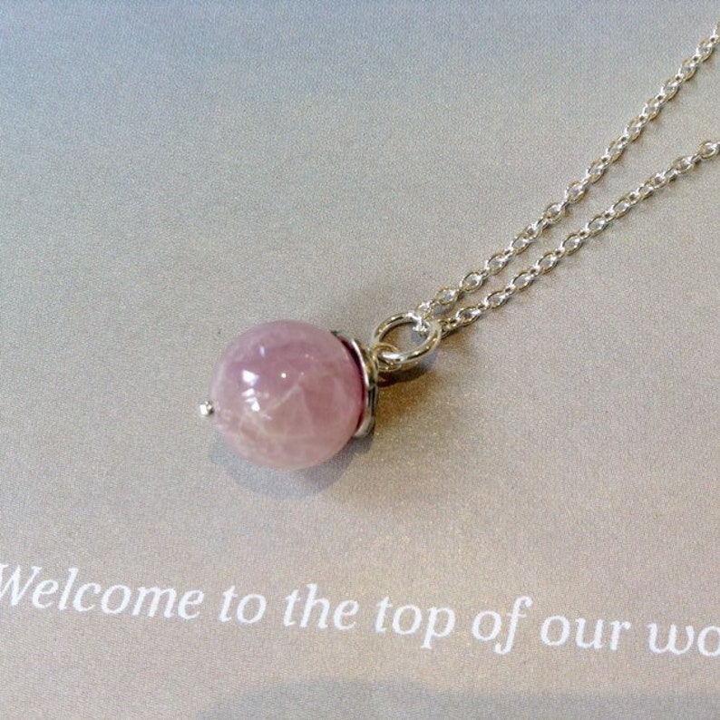 Op de afbeelding: Een delicate zilveren ketting met een ronde, lichtroze edelsteen hanger. De hanger wordt op zijn plaats gehouden door een zilveren zetting. De ketting is dun en de achtergrond is lichtgrijs. De tekst "Welcome to the top of our wo..." is zichtbaar.
