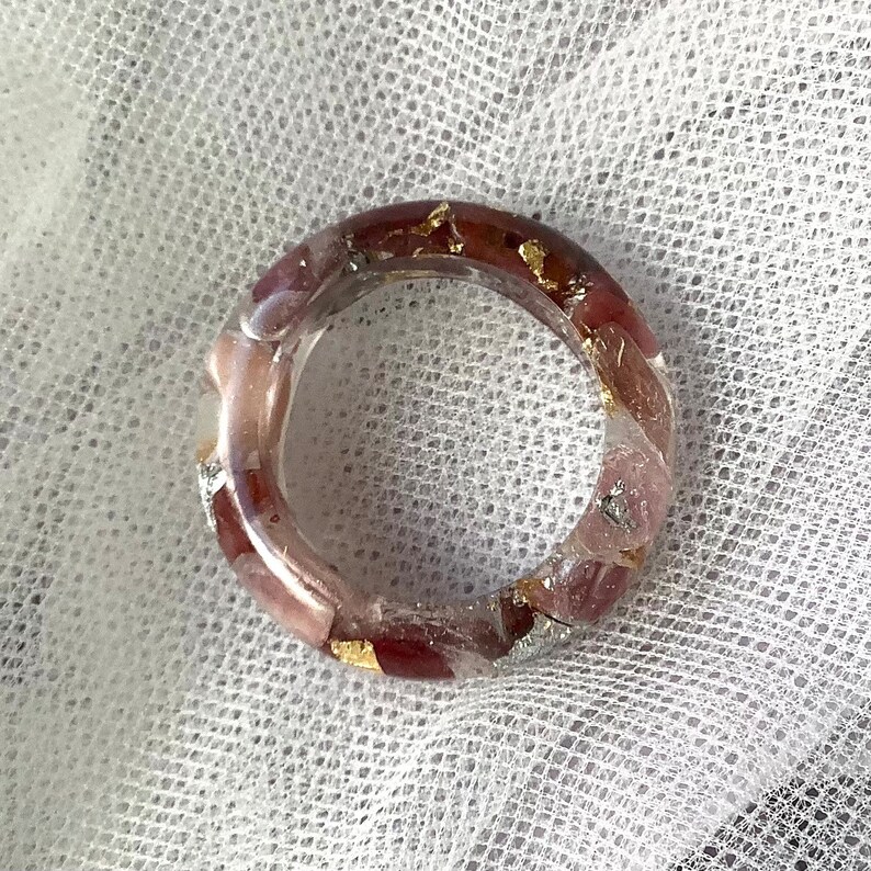 May include: A clear resin ring with embedded red and pink stones and gold leaf accents. The ring is set against a white, textured background. The ring is a unique piece of jewelry.