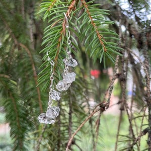 Peut inclure: Une boucle d'oreille en chaîne métallique argentée composée de quatre petites pierres de cristal transparentes et brutes suspendues en ligne verticale. La boucle est fixée à un crochet argenté et pend avec un aspect délicat et superposé.