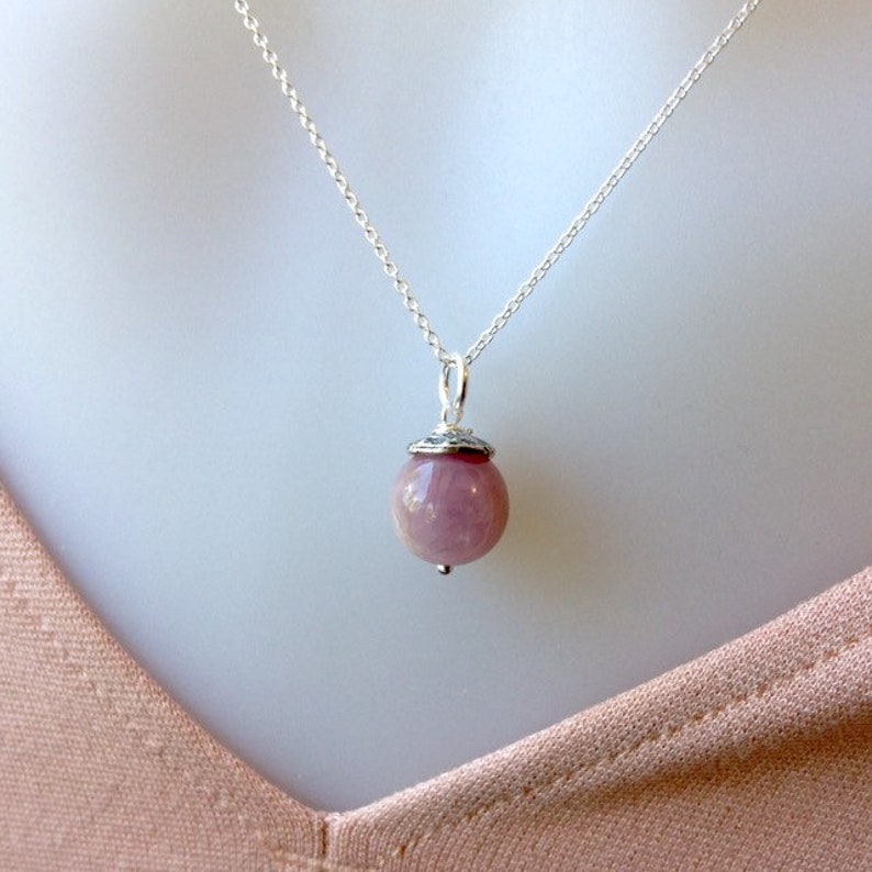 Op de afbeelding: Een zilveren ketting met een ronde, roze edelsteen hanger. De hanger heeft een zilveren dop en een kleine zilveren kraal. De ketting wordt getoond tegen een lichte achtergrond.