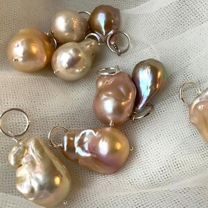 May include: A collection of irregularly shaped, iridescent pearls in shades of cream, peach, and mauve. Each pearl is attached to a small silver ring, suggesting they are intended for jewelry making or as pendants. The pearls are displayed on a white, textured surface.