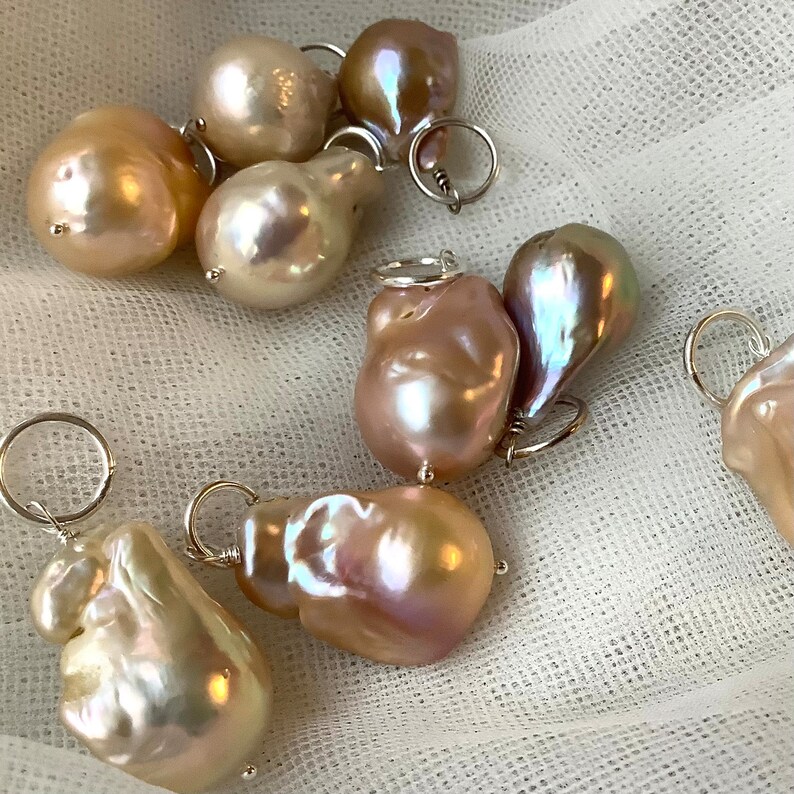 May include: A collection of irregularly shaped, iridescent pearls in shades of cream, peach, and mauve. Each pearl is attached to a small silver ring, suggesting they are intended for jewelry making or as pendants. The pearls are displayed on a white, textured surface.