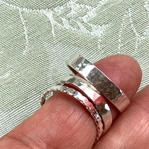 May include: A close-up of three silver rings. The top ring is a hammered band, the middle ring is a smooth band, and the bottom ring is a twisted rope design. The rings are displayed on a finger.