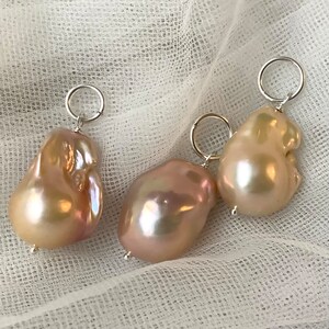 May include: Three baroque pearl pendants with silver-tone hoops. The pearls have a lustrous, iridescent sheen, displaying shades of pink, cream, and gold. The irregular shapes of the pearls add to their unique appeal. The pendants are displayed on a white, textured fabric.