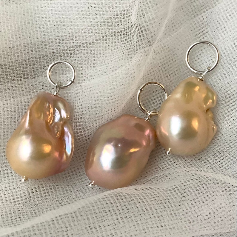 May include: Three baroque pearl pendants with silver-tone hoops. The pearls have a lustrous, iridescent sheen, displaying shades of pink, cream, and gold. The irregular shapes of the pearls add to their unique appeal. The pendants are displayed on a white, textured fabric.