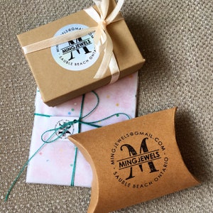 Op de afbeelding: Drie sieradendoosjes in verschillende maten en vormen, verpakt en klaar om cadeau te geven. De dozen zijn bruin en vastgebonden met linten en touw. De dozen hebben de tekst "MING JEWELS" en het e-mailadres "MINGJEWELS@GMAIL.COM" erop gedrukt.