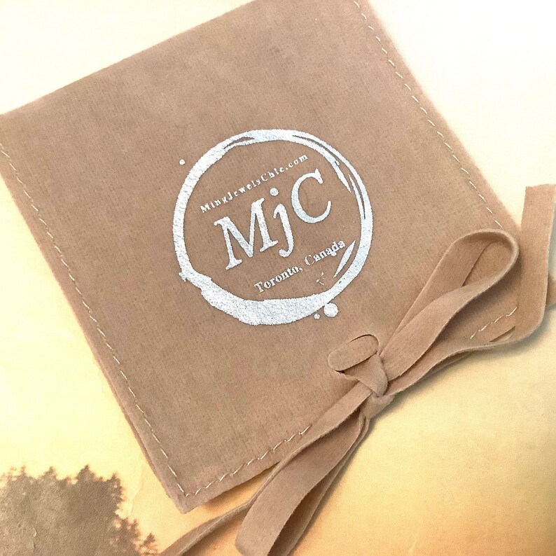 May include: A tan-colored velvet pouch with a tied ribbon closure. The pouch features a silver-colored circular logo with the text "MjC" and "MingJewelsChic.com Toronto, Canada."
