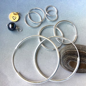 May include: A collection of silver hoop earrings in different sizes, a small gold pendant with a heart, and a dark pearl earring. The jewellery is displayed on a light grey surface, with a dark stone in the background.