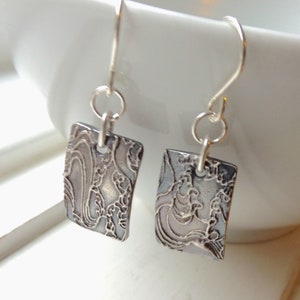 Peut inclure: Une paire de boucles d'oreilles en argent avec un motif rectangulaire texturé. Les boucles d'oreilles présentent un motif tourbillonnant détaillé en argent plus foncé et sont suspendues à des crochets en argent. Elles sont présentées sur fond blanc.