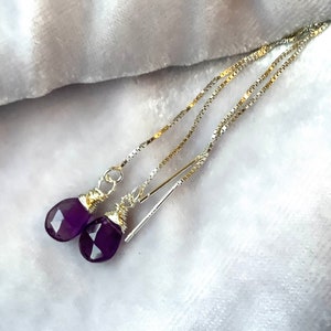 May include: A pair of amethyst drop earrings. Each earring has a faceted, deep purple gemstone hanging from a delicate silver chain. The gemstones are wire-wrapped in gold-coloured metal.