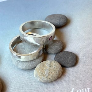 May include: Two silver rings with a hammered texture are stacked, one with a small pink gemstone. The rings rest on smooth grey stones. The image presents a natural and elegant display of the jewellery.