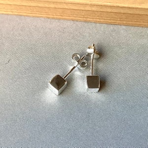 May include: A pair of small, square silver stud earrings. Each earring has a geometric cube design. The earrings are displayed on a light blue surface, with a wooden background.