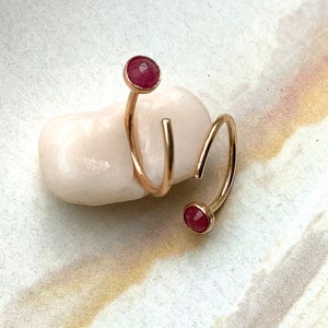 May include: A gold ring with a spiral design, featuring two round, red gemstones. The ring is displayed on a white, rounded stone, showcasing its elegant and modern style. The gemstones are a deep red colour.