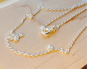 May include: A delicate silver necklace with a classic cable chain design. The necklace features a lobster clasp closure and is displayed on a light brown wooden surface. The chain is approximately 45 cm long.