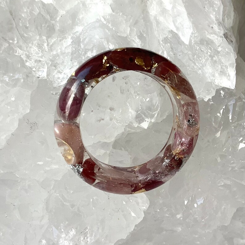 May include: A clear resin ring with embedded burgundy and pink stones, accented with gold and silver flakes. The ring has a smooth, polished finish, showcasing the natural elements within. A unique piece of jewelry.