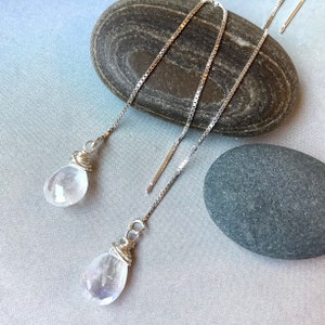 May include: Silver threader earrings featuring clear, teardrop-shaped gemstones. The gemstones are wrapped in silver wire. The earrings are presented on a light grey surface with two grey stones in the background.