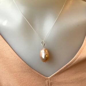 May include: A silver necklace with a large, teardrop-shaped, peach-colored pearl pendant. The pearl has a smooth, lustrous surface and hangs from a small silver loop. The chain is delicate and hangs on a neutral background.