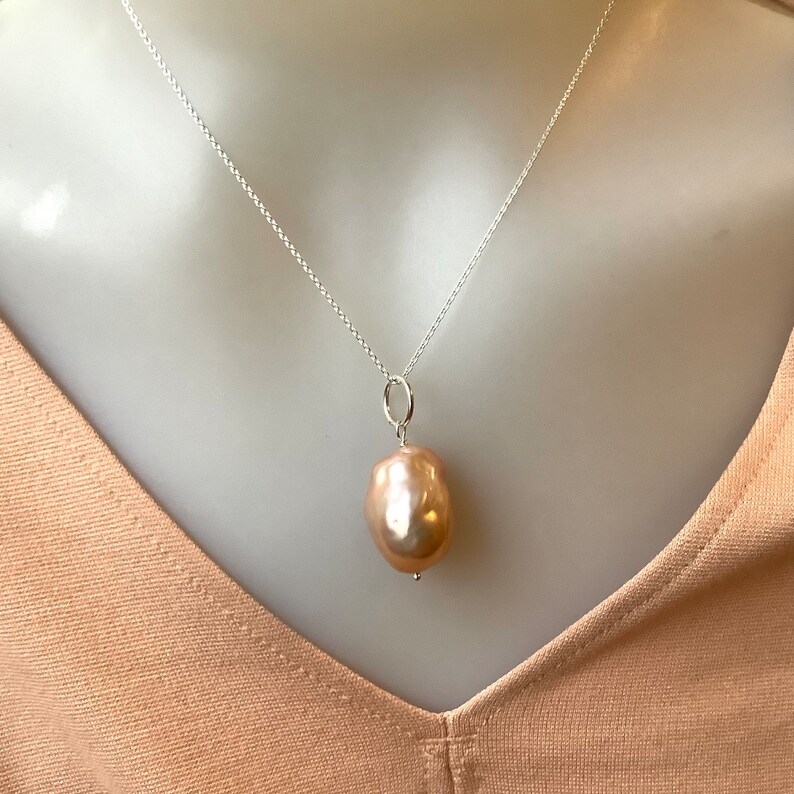 May include: A silver necklace with a large, teardrop-shaped, peach-colored pearl pendant. The pearl has a smooth, lustrous surface and hangs from a small silver loop. The chain is delicate and hangs on a neutral background.