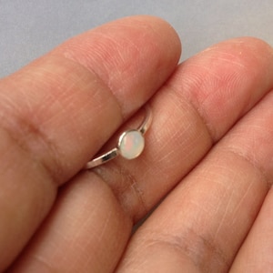 May include: A delicate silver ring with a small, round, iridescent opal gemstone. The opal has a milky white base with subtle flashes of colour. The ring's band is thin and polished, creating a minimalist and elegant design.