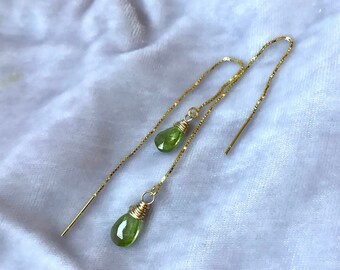 May include: Elegant gold threader earrings with pear-shaped, light green peridot gemstones. The peridot stones are wire-wrapped and hang from long, thin gold chains. The earrings are shown on a white, textured surface.