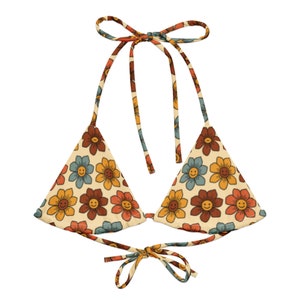 May include: A retro-style bikini top with a floral pattern. The top features a cream-colored background with smiling flowers in shades of brown, yellow, and blue. The halter top has tie closures at the neck and back, and is a triangle shape.