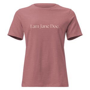 以下が含まれることがあります： 胸の中央に白い文字で「I am Jane Doe.」とプリントされた、ダスティローズカラーの半袖Tシャツ。クルーネックで標準的なフィット感があり、控えめな質感の無地生地で作られています。