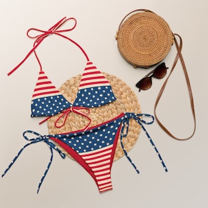 May include: A red, white, and blue bikini with a star-spangled pattern. The bikini is laid out on a woven straw mat with a straw bag and sunglasses.