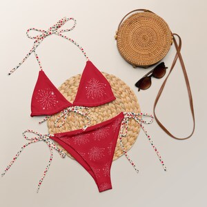 May include: Red bikini with a white firework pattern. The bikini top has adjustable straps and ties at the back. The bikini bottom has ties on both sides. The bikini is on a straw mat with a round straw bag and sunglasses.
