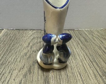 May include: A small, white ceramic vase with a heart-shaped opening and blue trim. Two small figurines of children in blue and white outfits are kissing at the base of the vase. The vase has blue floral accents.