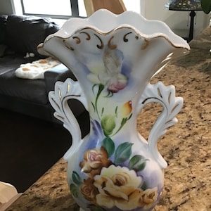 May include: A white porcelain vase with gold accents and a floral design. The vase has two handles and a scalloped rim. The flowers are painted in shades of yellow, pink, and brown.