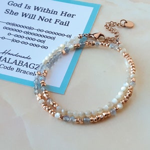 Könnte beinhalten: Ein doppelreihiges Armband mit weißen und blauen Perlen und einem roségoldenen Verschluss. Das Armband liegt auf einer weißen Oberfläche mit einer Karte, auf der "God Is Within Her She Will Not Fail" und "Handmade by MALABAGZ Morse Code Bracelet" steht.