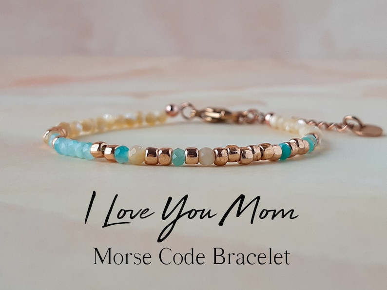 May include: A delicate bracelet with a message in Morse code. The bracelet is made with small, light blue, white, and gold beads. The message on the bracelet reads "I Love You Mom". Morse Code Bracelet.