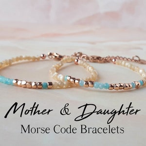 Puede incluir: Dos pulseras de cuentas, una más grande y otra más pequeña, con un esquema de color beige claro y turquesa. Las pulseras están hechas con cuentas pequeñas y tienen un cierre de oro rosa. El texto "Pulseras de código Morse madre e hija" se muestra debajo de las pulseras.
