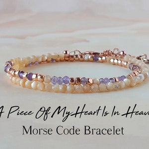 Puede incluir: Un brazalete de doble cadena con un diseño delicado. El brazalete presenta pequeñas cuentas de color claro y cuentas moradas, intercaladas con cuentas doradas. El brazalete está hecho con un cierre de oro rosa y tiene un dije que dice "A Piece Of My Heart Is In Heaven" en escritura cursiva. Brazalete de código Morse.