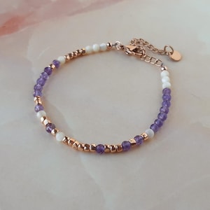 Op de afbeelding: Een delicate armband met paarse en witte kralen, afgewisseld met goudkleurige kralen. De armband heeft een karabijn sluiting.