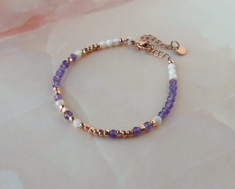 Op de afbeelding: Een delicate armband met paarse en witte kralen, afgewisseld met goudkleurige kralen. De armband heeft een karabijn sluiting.