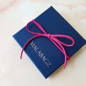 May include: A navy blue gift box with a pink ribbon tied around it. The box has the text "MALABAGZ" printed on the front in gold lettering.