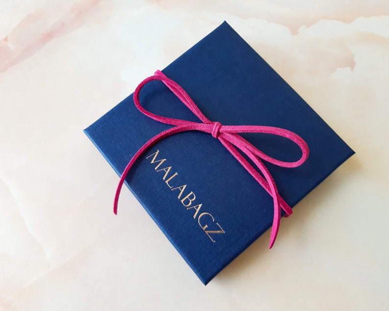 May include: A navy blue gift box with a pink ribbon tied around it. The box has the text "MALABAGZ" printed on the front in gold lettering.